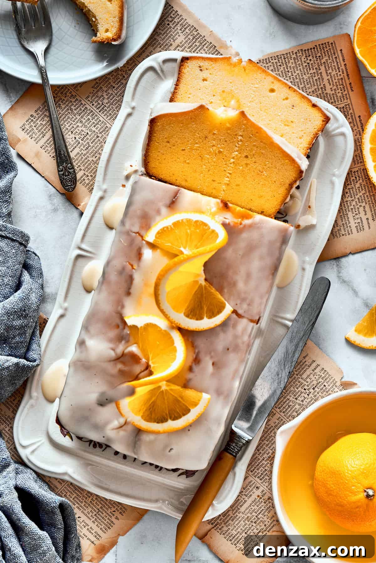 A sliced orange loaf cake topped with orange glaze, orange slices, and wish more oranges surrounding it.