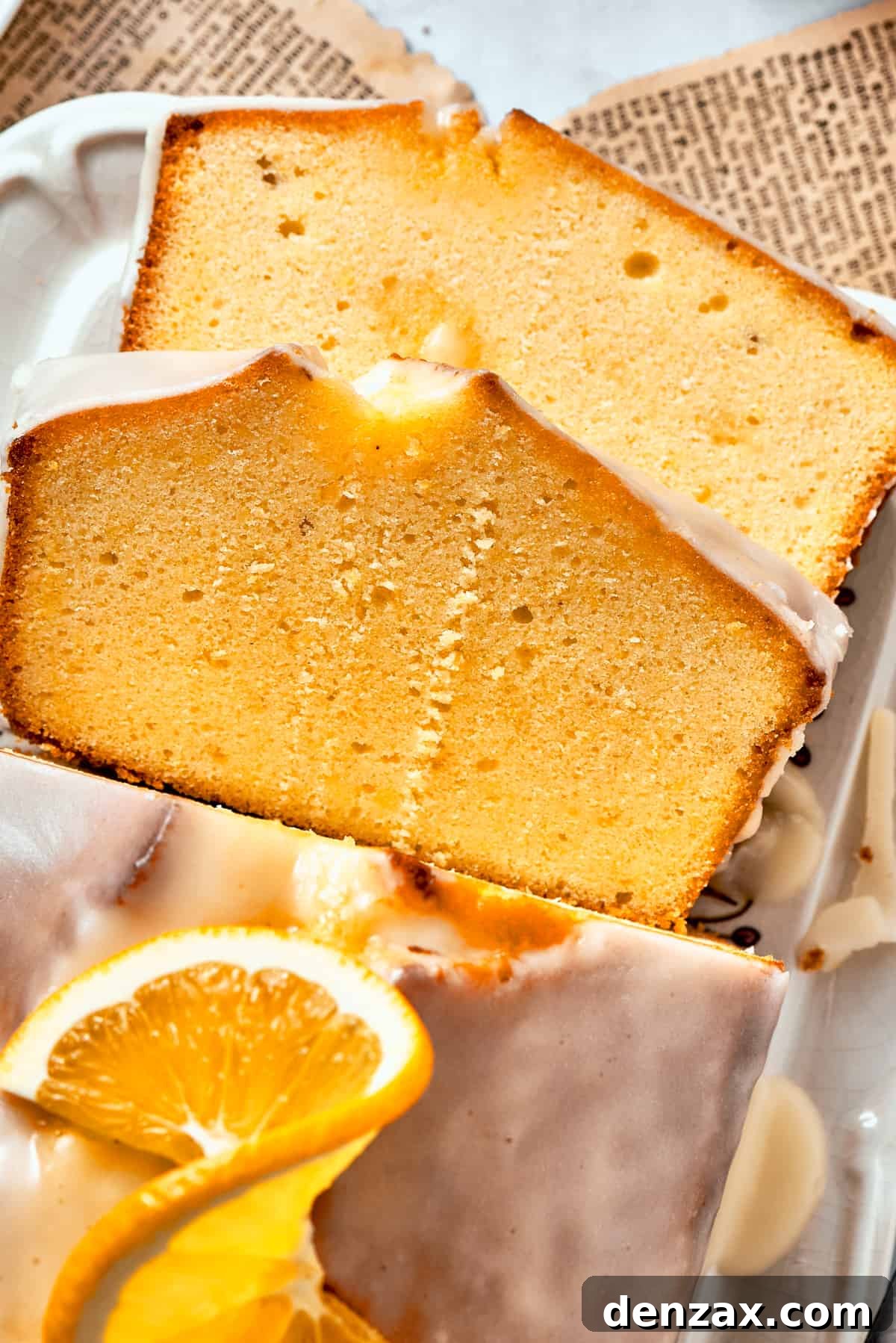 Two slices of glazed orange cake rest atop each other, with the rest of the loaf visible nearby topped with orange slices.