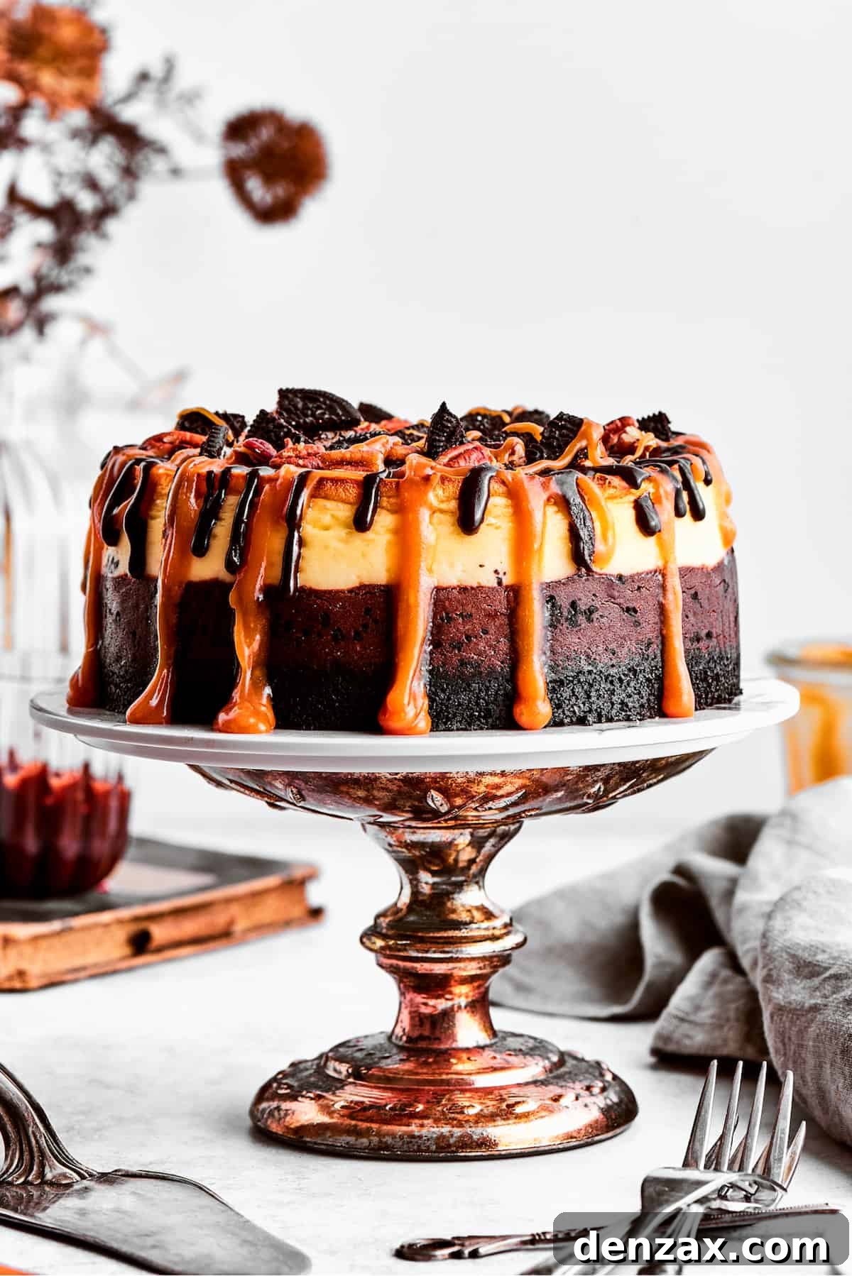 A turtle cheesecake dripping with caramel sauce is displayed on a bronze cake stand, ready to be sliced and served.