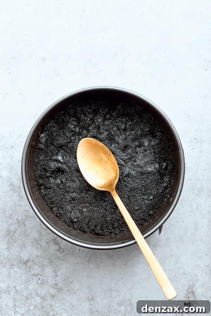 A spoon pats a chocolate cookie crust evenly into a springform baking pan, creating a firm base for the cheesecake.
