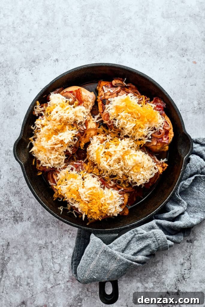 Four chicken breasts are topped with bacon and cheese in a cast iron skillet, ready for baking.