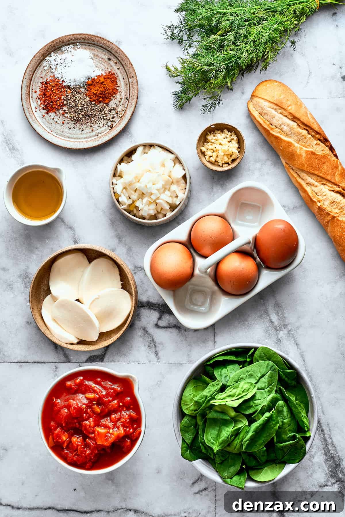 All the fresh ingredients for making Eggs in Purgatory are neatly arranged on a white background: fresh eggs, a loaf of rustic bread, fresh mozzarella cheese, a pile of baby spinach, canned diced tomatoes, garlic cloves, olive oil, and a selection of spices including red pepper flakes and dill.
