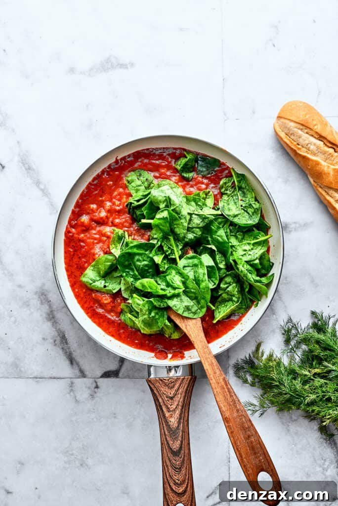 Vibrant green baby spinach leaves are actively stirred into the simmering homemade red tomato sauce within a skillet, adding freshness and nutrients.