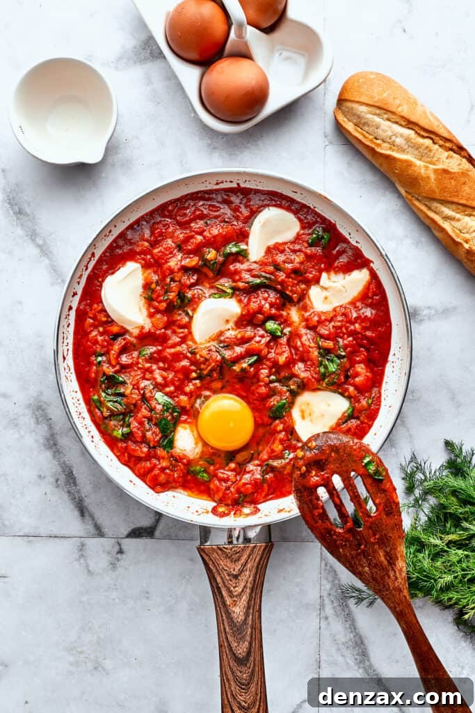 Four fresh eggs have just been cracked and carefully placed into individual divots within a rich skillet of tomato sauce, spinach, and melting mozzarella cheese.
