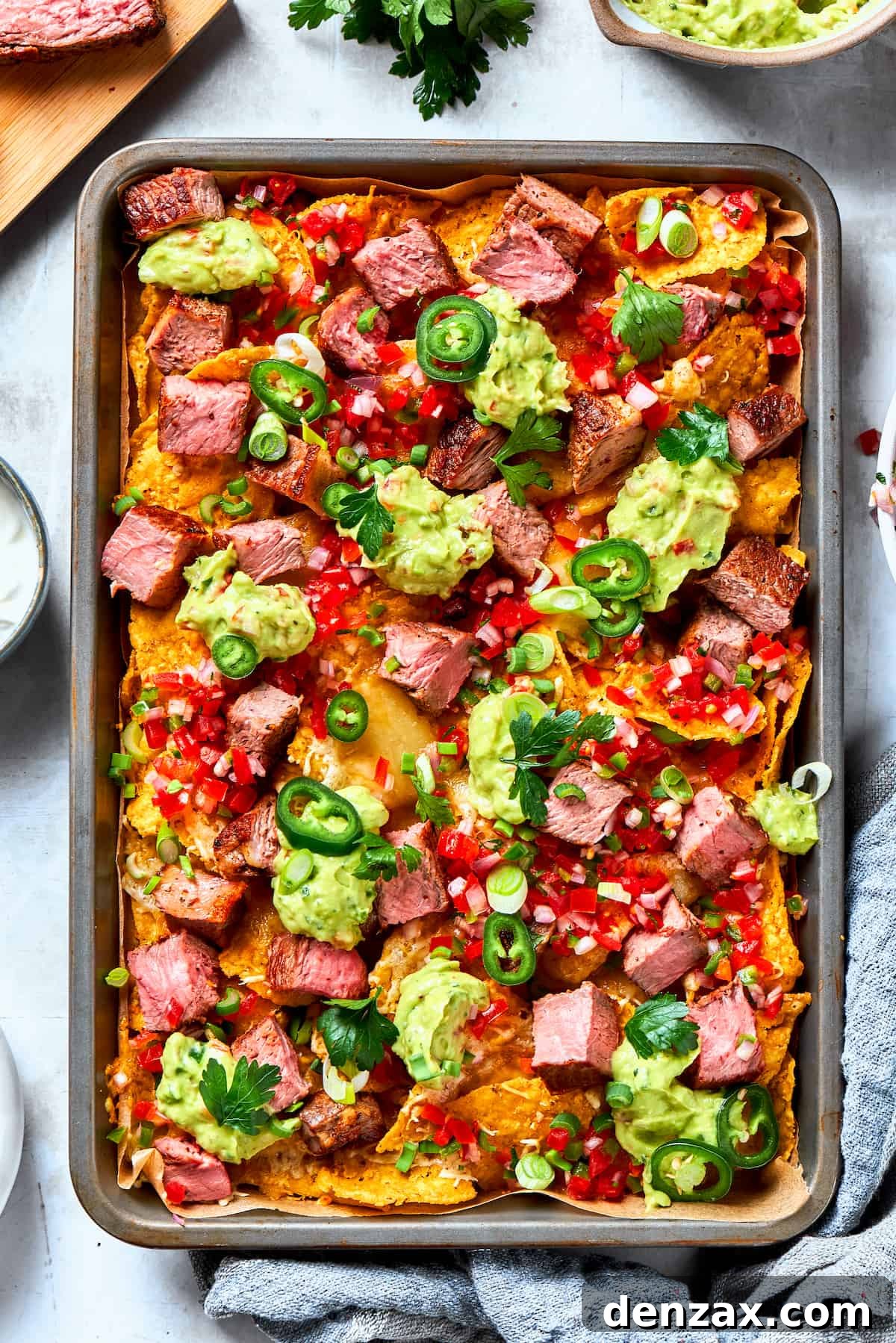 A baked pan of steak nachos is topped with dollops of guacamole and chunks of steak.