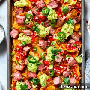 A baked pan of steak nachos is topped with dollops of guacamole and chunks of steak.