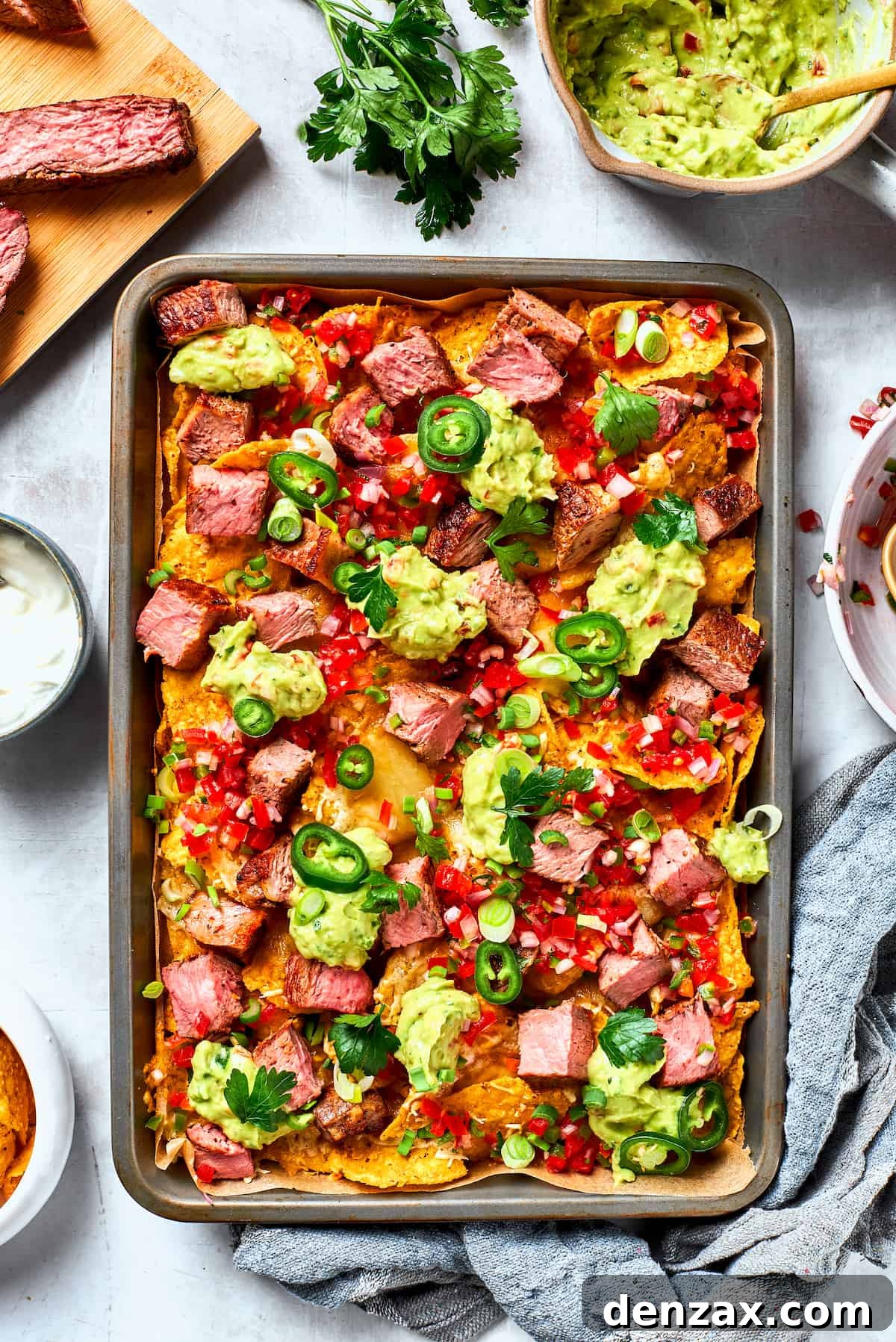 A metal baking sheet of steak nachos is surrounded by ore steak and toppings.