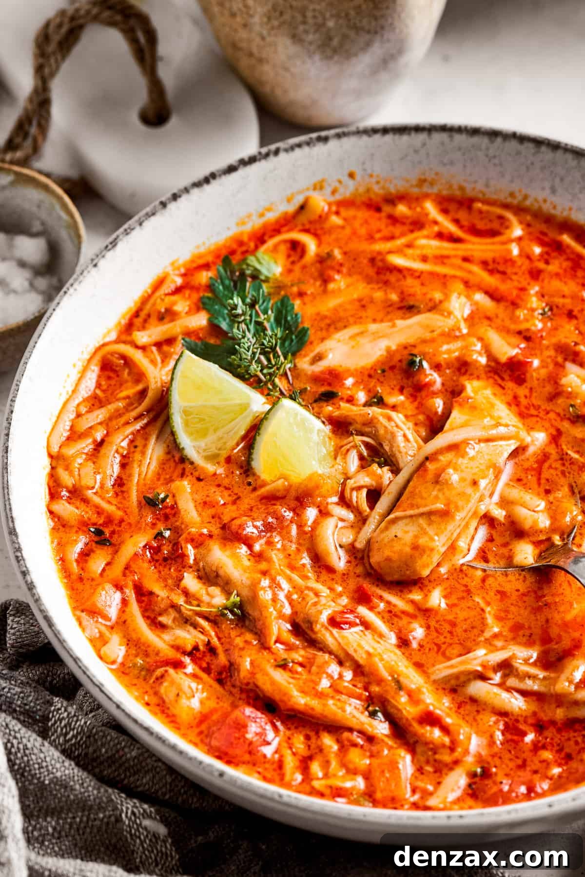 Close-up view of a single bowl of spicy chicken soup, garnished with fresh cilantro and lime wedges, with a spoon resting in it.