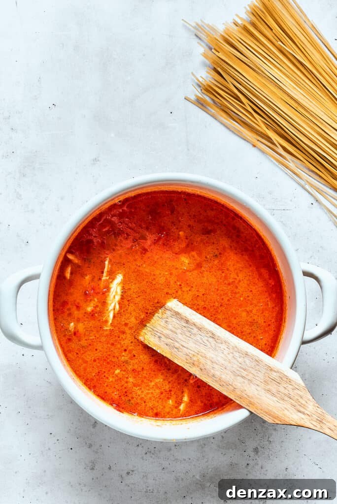 Delicate rice noodles are being carefully added to a simmering pot of spicy chicken soup, with more noodles prepared nearby.