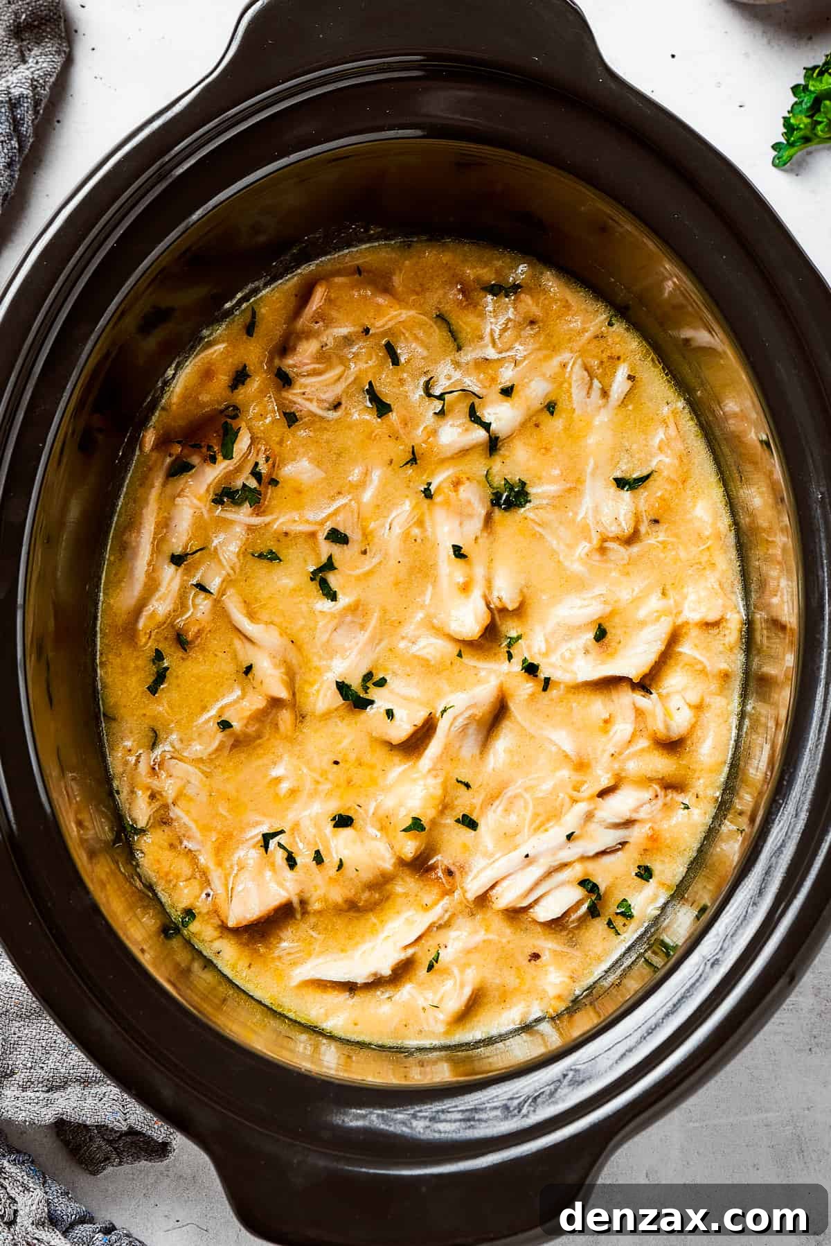 Overhead image of Shredded chicken in a creamy sauce cooking in a black slow cooker, garnished with fresh parsley.