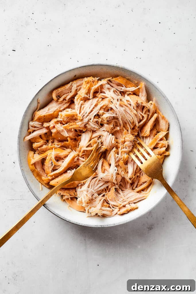 Cooked chicken is being expertly shredded into tender pieces using two forks in a white serving bowl, a key step for mixing back into the creamy gravy.