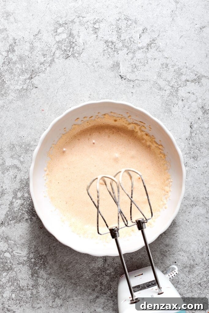 A hand mixer is poised to mix the warmed egg mixture in a bowl until it is thick and cooled.