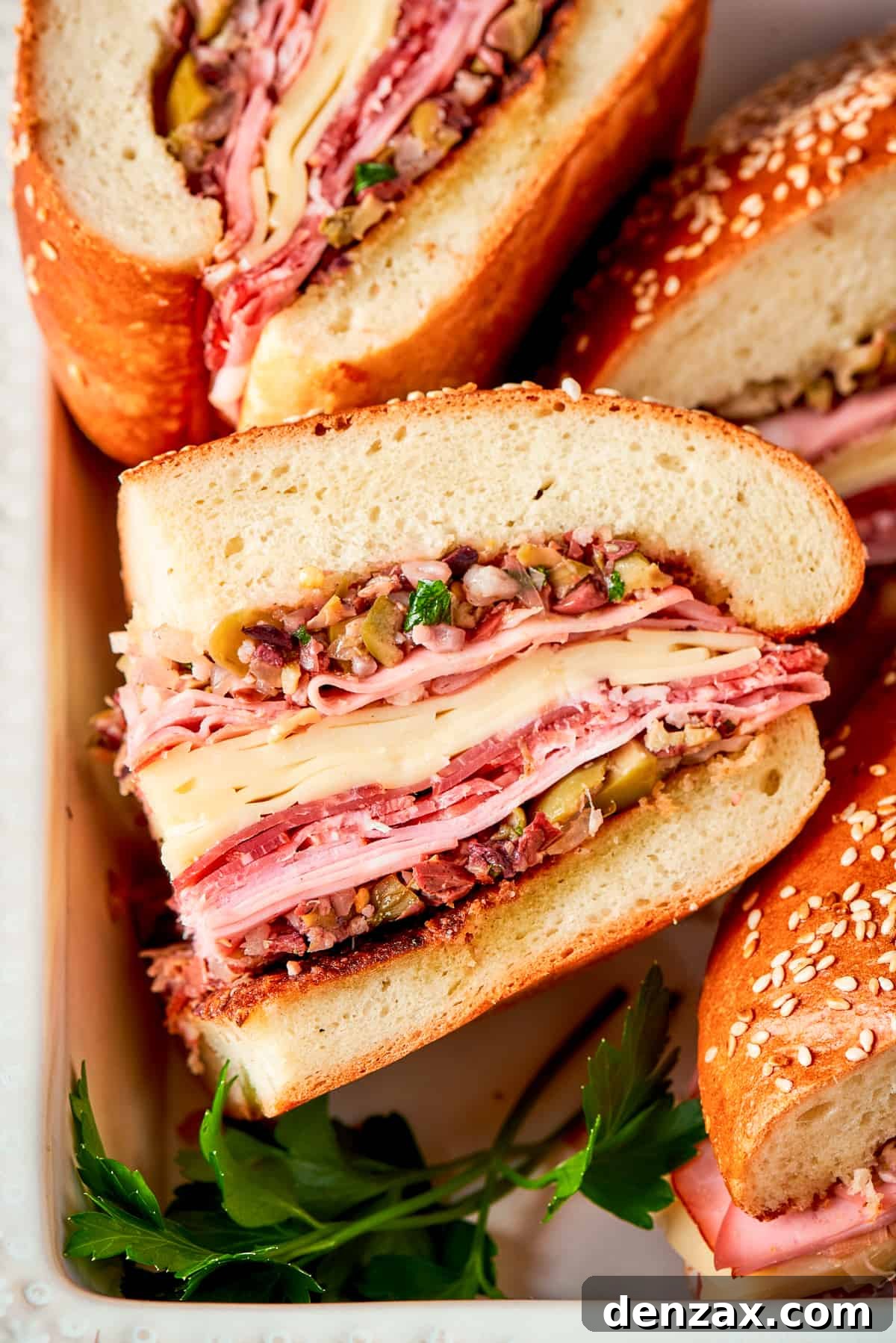 A towering Muffaletta sandwich, sliced in half to reveal abundant layers of homemade olive salad, creamy provolone and swiss cheeses, and a variety of Italian deli meats on soft sesame-seeded bread.
