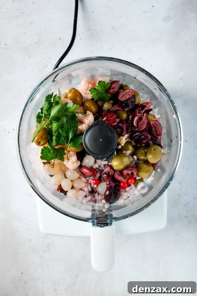 All the ingredients for the olive salad, including various olives, pickled vegetables, and fresh herbs, are being added to a food processor.