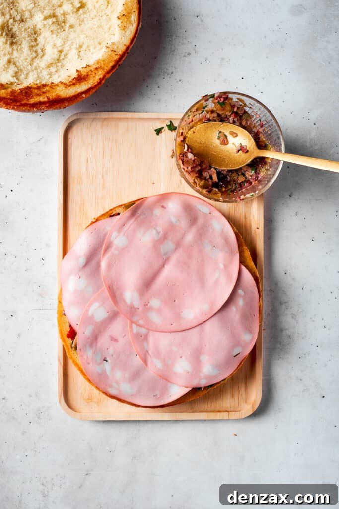The final slices of Mortadella are carefully arranged on top of other deli meats, forming a thick, flavorful layer for the muffaletta sandwich.