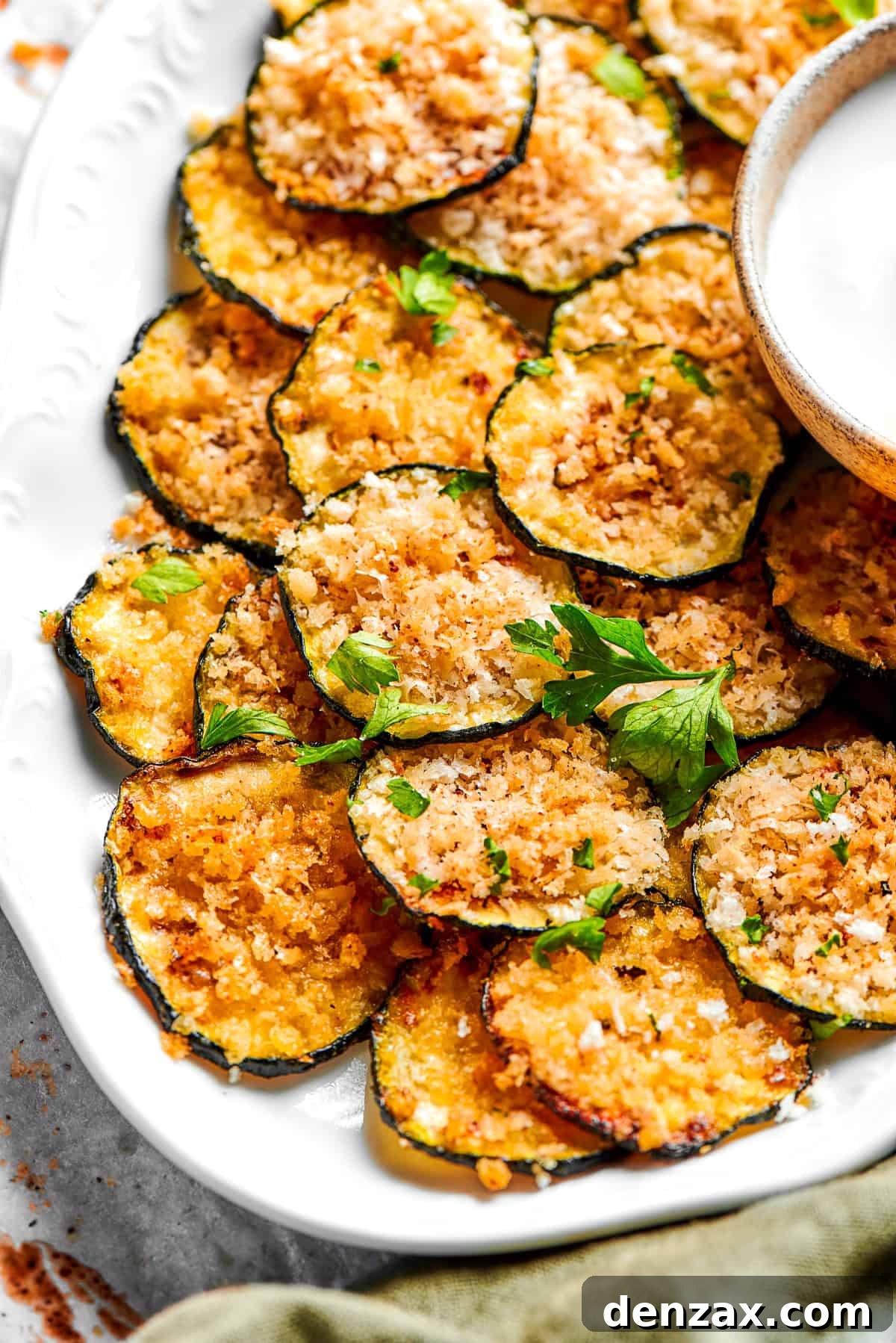 A pile of golden, crispy zucchini chips arranged enticingly on a white plate, garnished with fresh herbs.