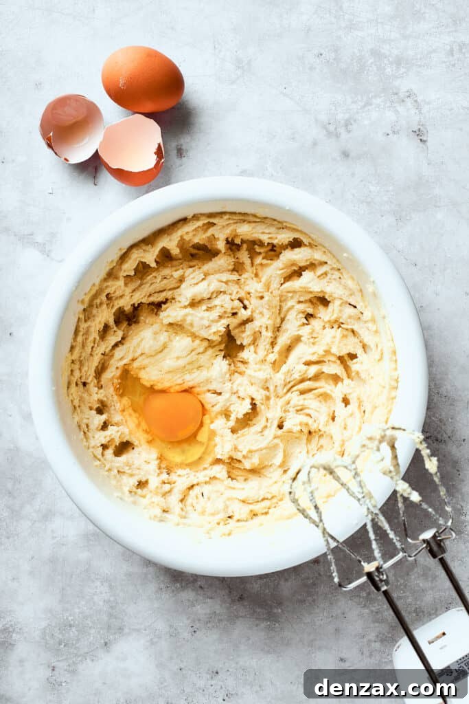 A cracked egg is being added to the creamy butter and sugar mixture in a bowl, ready to be beaten in.