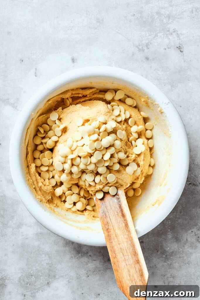 Sweet white chocolate chips are being gently folded into the prepared cookie dough in a mixing bowl.