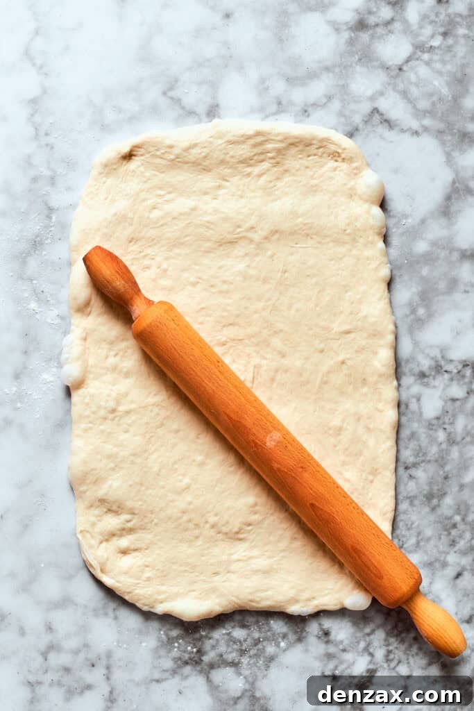 Pizza dough being rolled out evenly with a rolling pin on a lightly floured surface.
