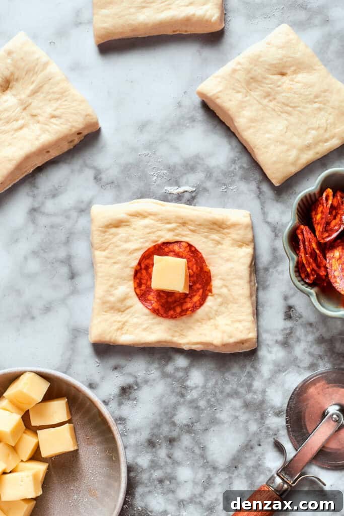 A square of pizza dough with a slice of pepperoni and a cube of mozzarella cheese placed precisely in the center, ready for folding.
