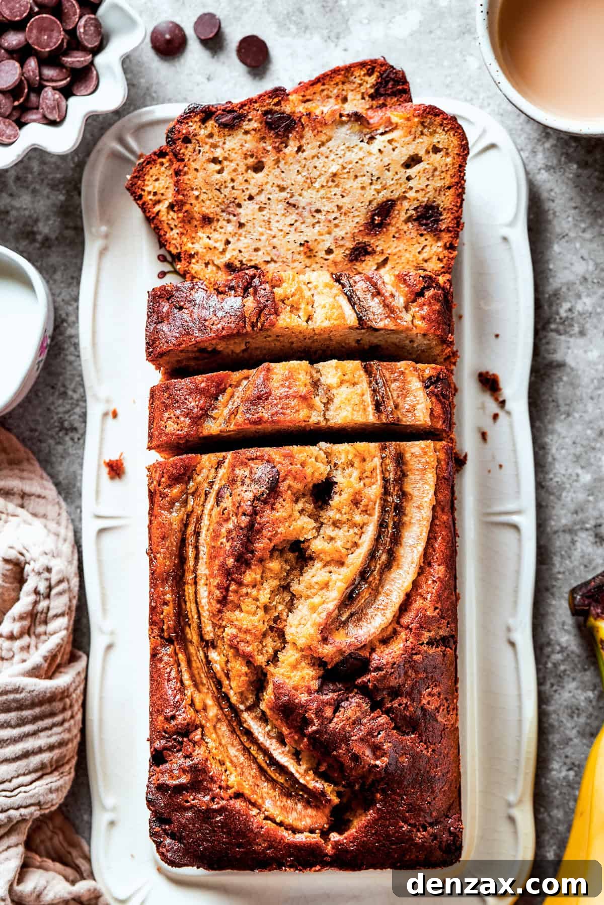 A perfectly baked loaf of Sour Cream Banana Bread, adorned with chocolate chips and a caramelized banana slice on top, resting in a loaf pan, surrounded by fresh bananas and other baking ingredients.