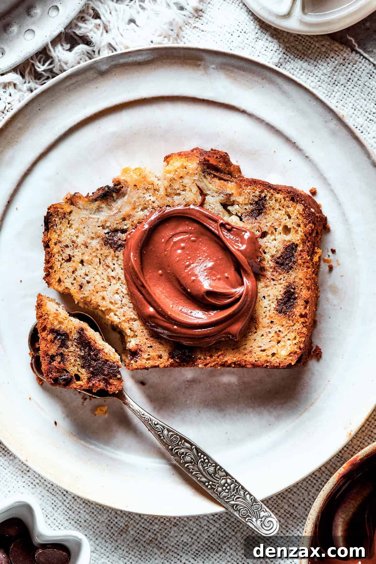 A hand is delicately spreading a generous layer of creamy Nutella over a thick slice of freshly baked Sour Cream Banana Bread, highlighting its moist texture and inviting appeal.