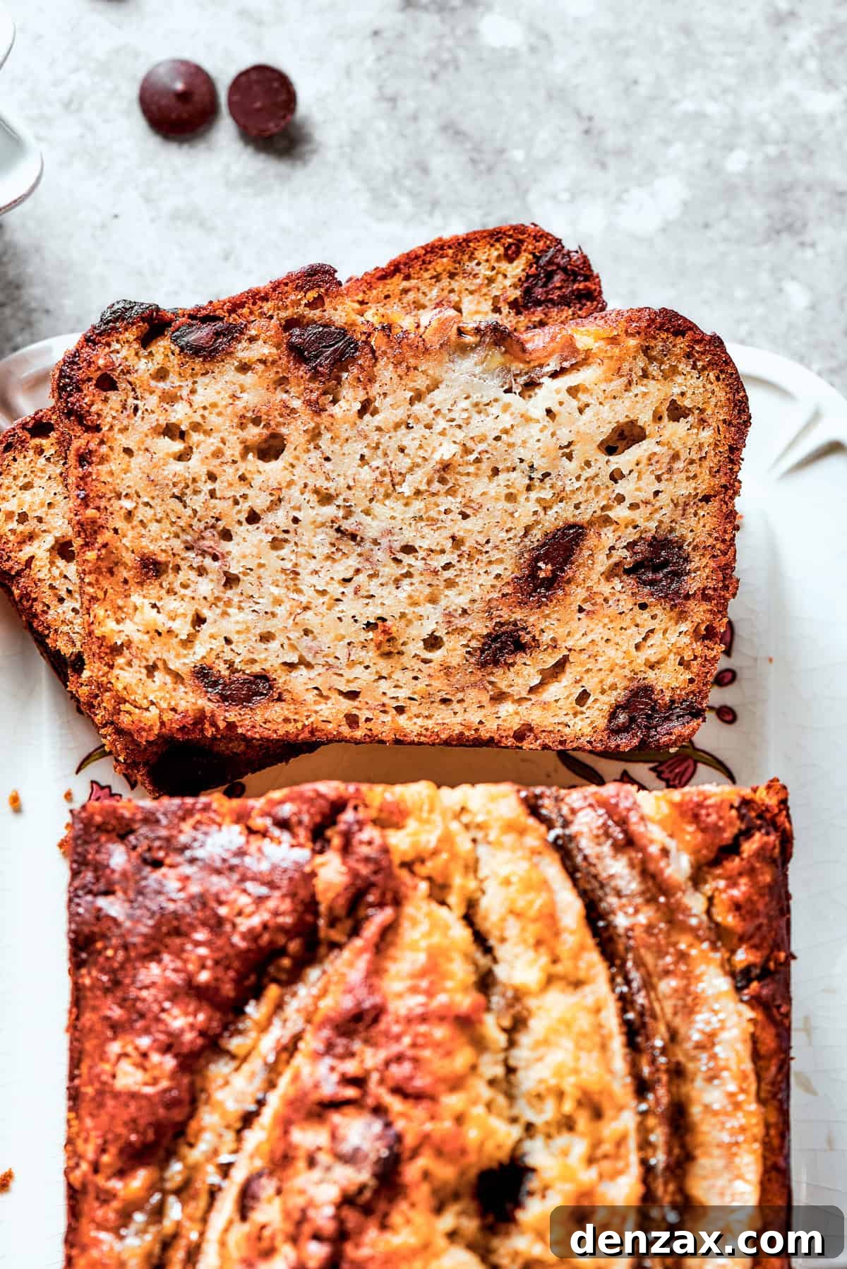 An inviting overhead view of a freshly baked Sour Cream Banana Bread loaf, with two slices cut out, revealing its moist, tender interior studded with chocolate chips. The remaining loaf rests on a cooling rack.