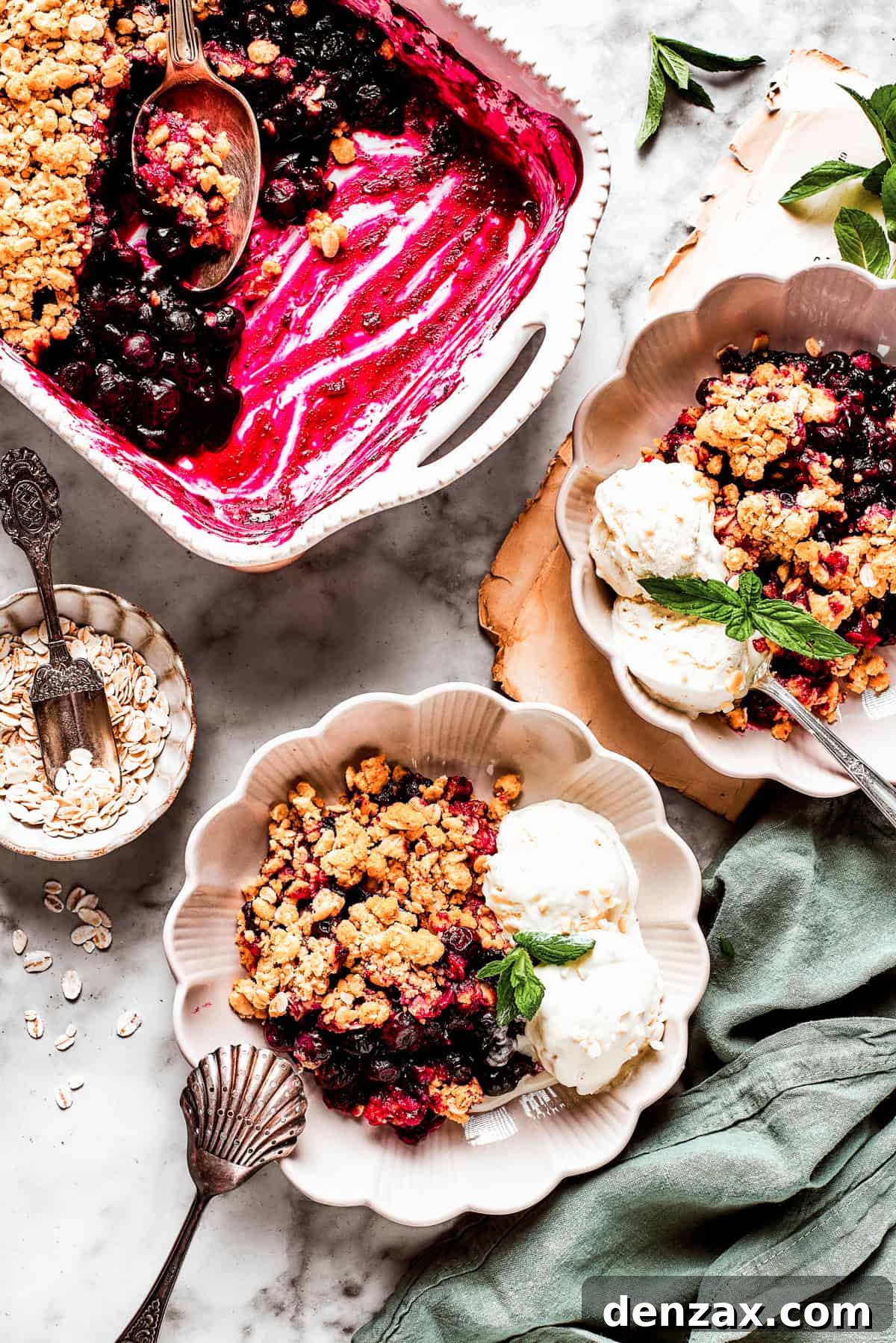 Overhead image of two dessert bowls of blueberry crisp served with scoops of ice cream. There is a baking dish with the crisp placed near the bowls.