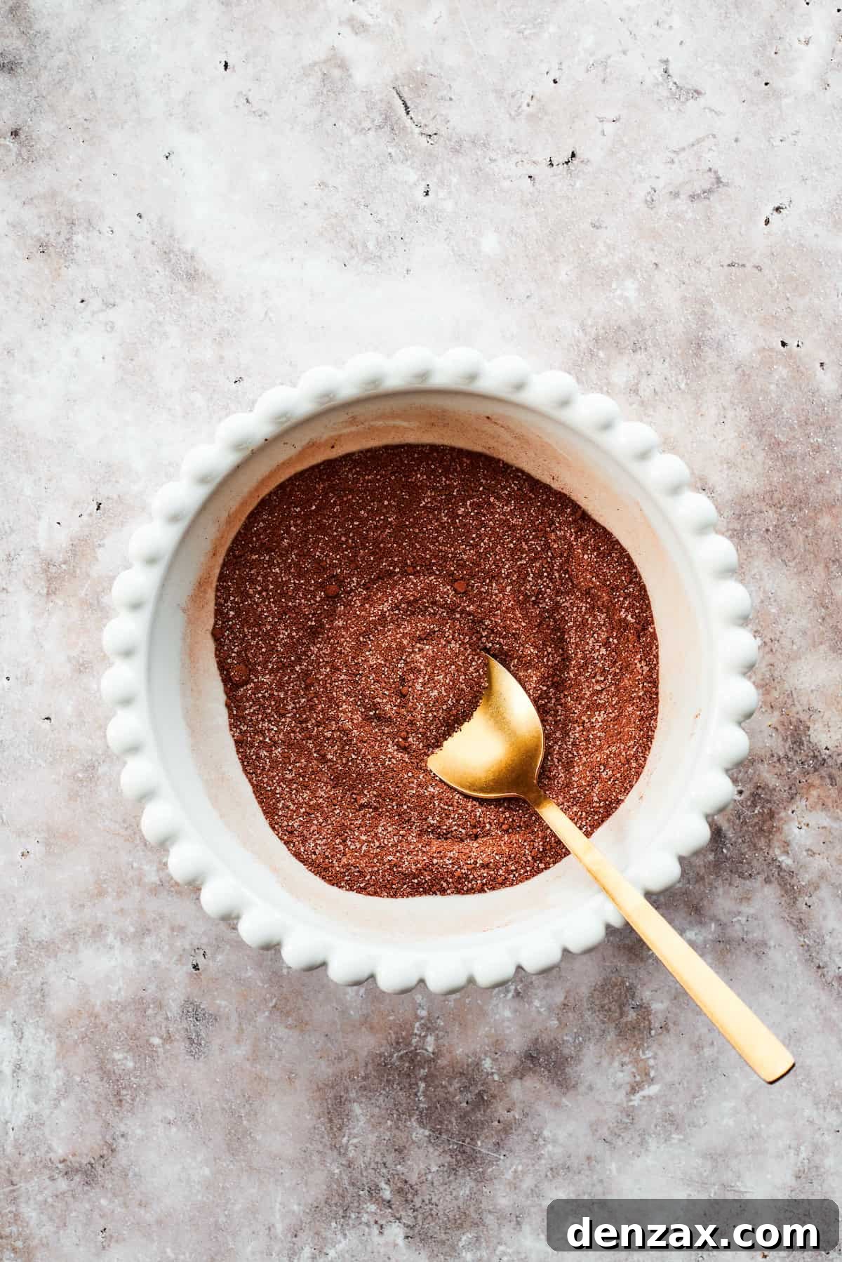 A spoon meticulously mixes the dry fudge topping ingredients in a small bowl, ensuring they are well combined.