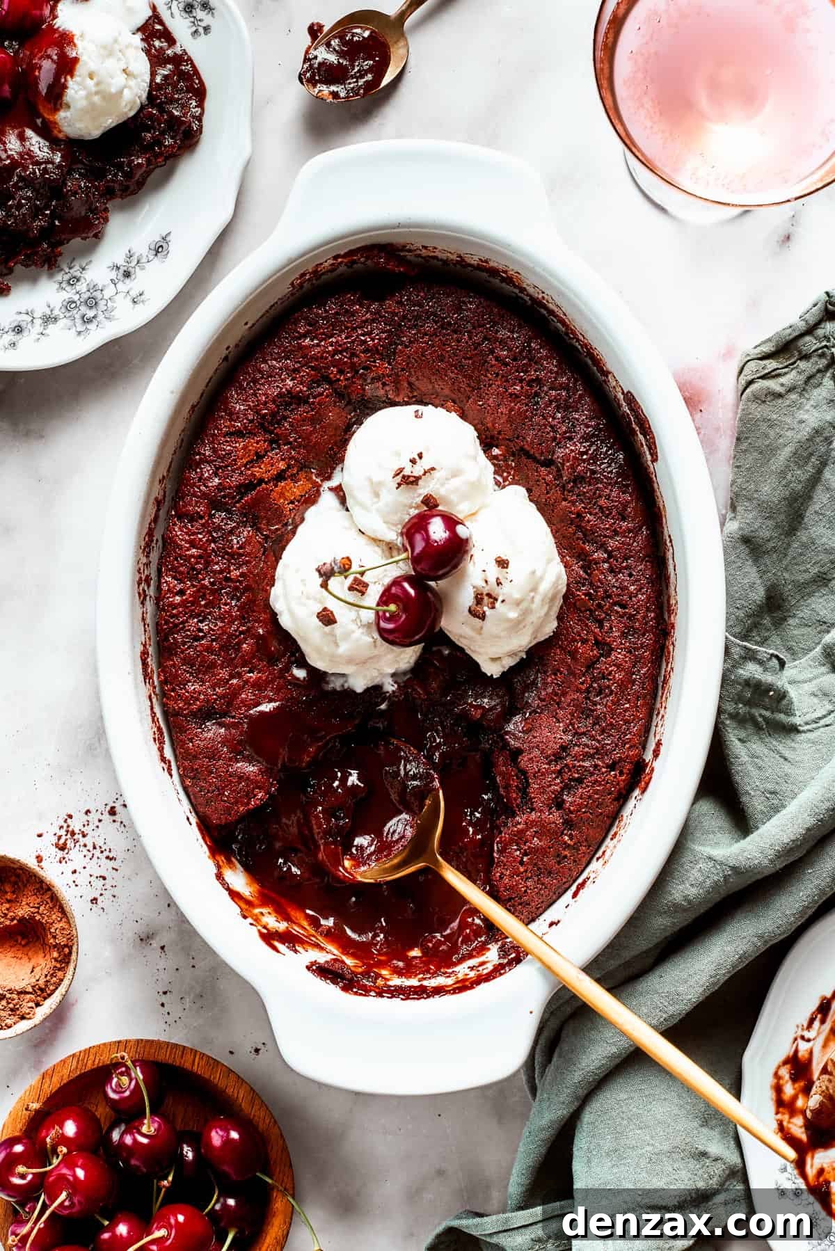 A warm chocolate pudding cake in a casserole dish, beautifully garnished with scoops of vanilla ice cream and two vibrant cherries, with a gold spoon poised to serve.