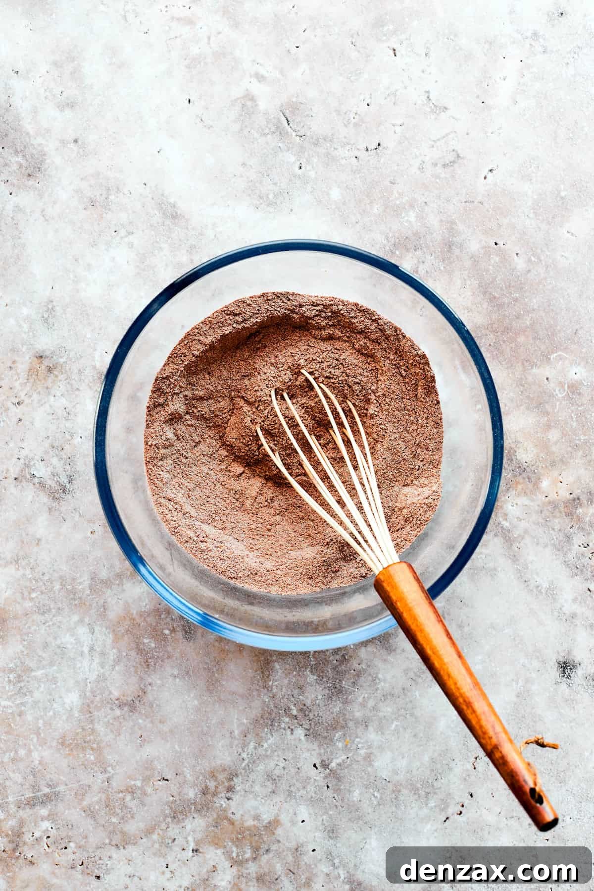 The dry ingredients for the chocolate pudding cake, comprising flour, sugars, cocoa, and baking powder, are being whisked thoroughly in a bowl.