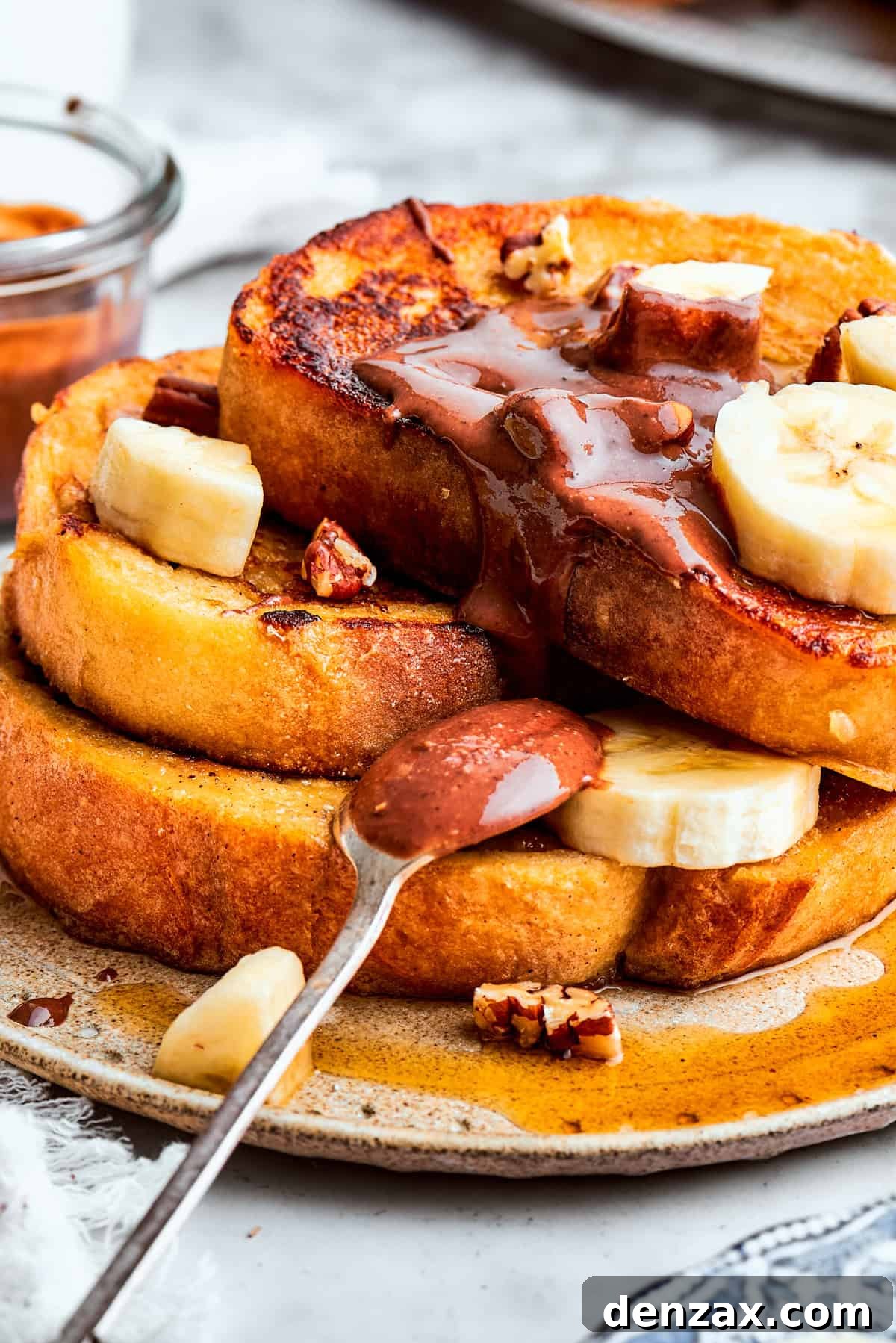 A close-up shot of a magnificent stack of Banana French Toast, artfully garnished with slices of fresh banana, a dusting of powdered sugar, and a generous drizzle of luscious chocolate hazelnut spread, showcasing its irresistible texture.