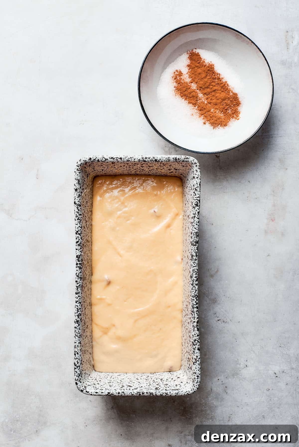 Amish friendship bread batter filling a loaf pan, with a small bowl of cinnamon sugar topping beside it.