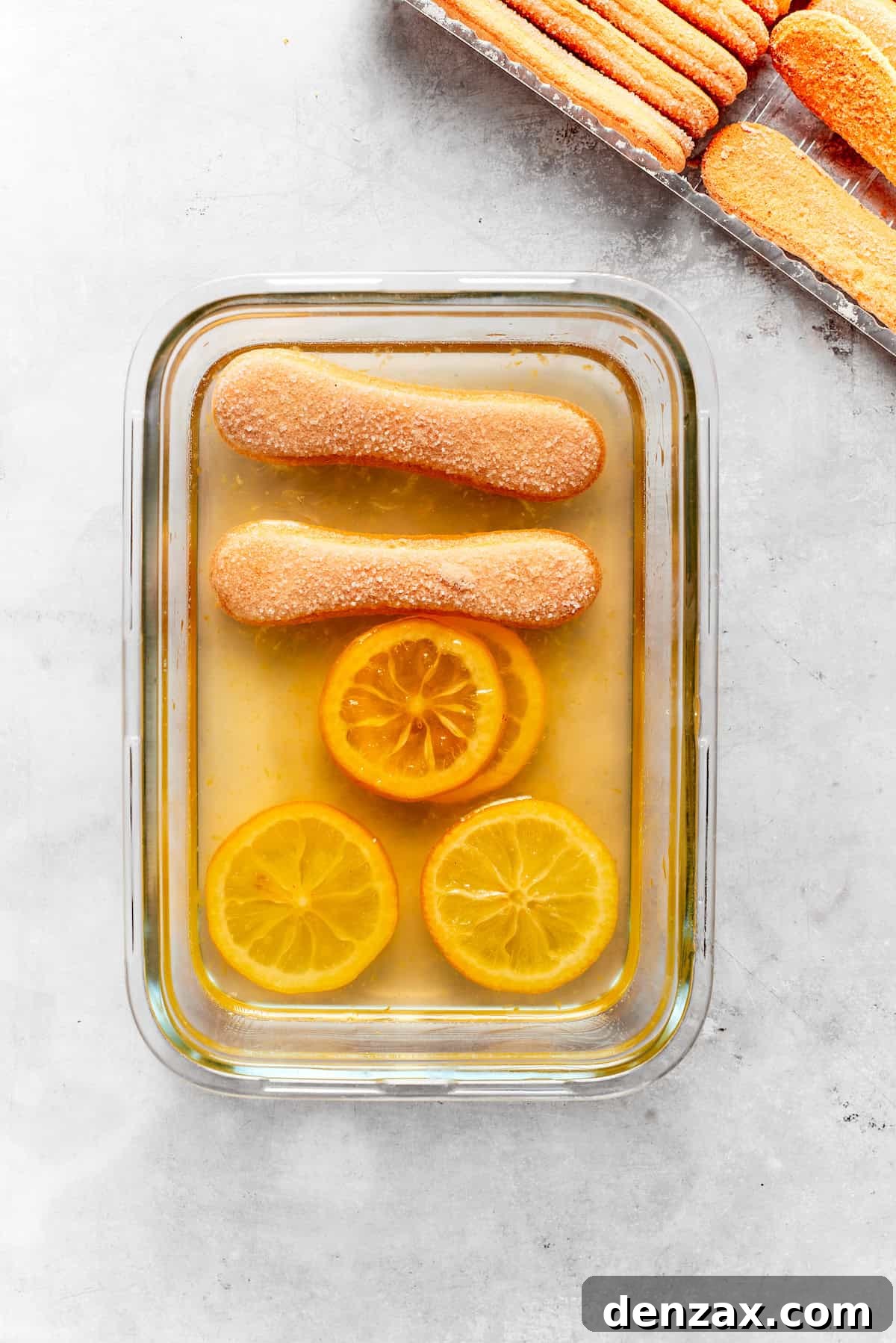 Ladyfinger cookies, briefly dipped in lemon syrup, are meticulously arranged in a single layer at the bottom of a baking dish.