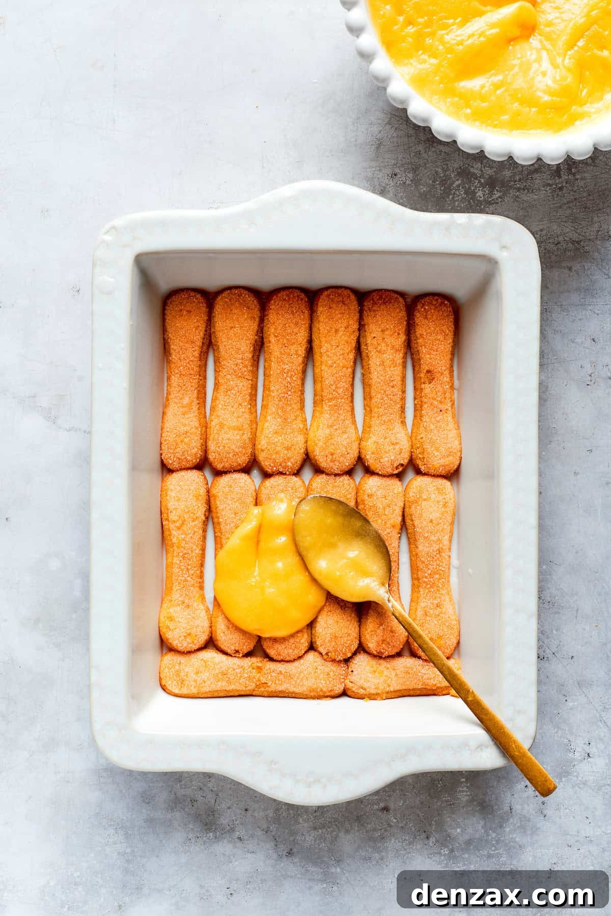 A generous layer of smooth, golden lemon curd is evenly spread over the arranged ladyfinger cookies.