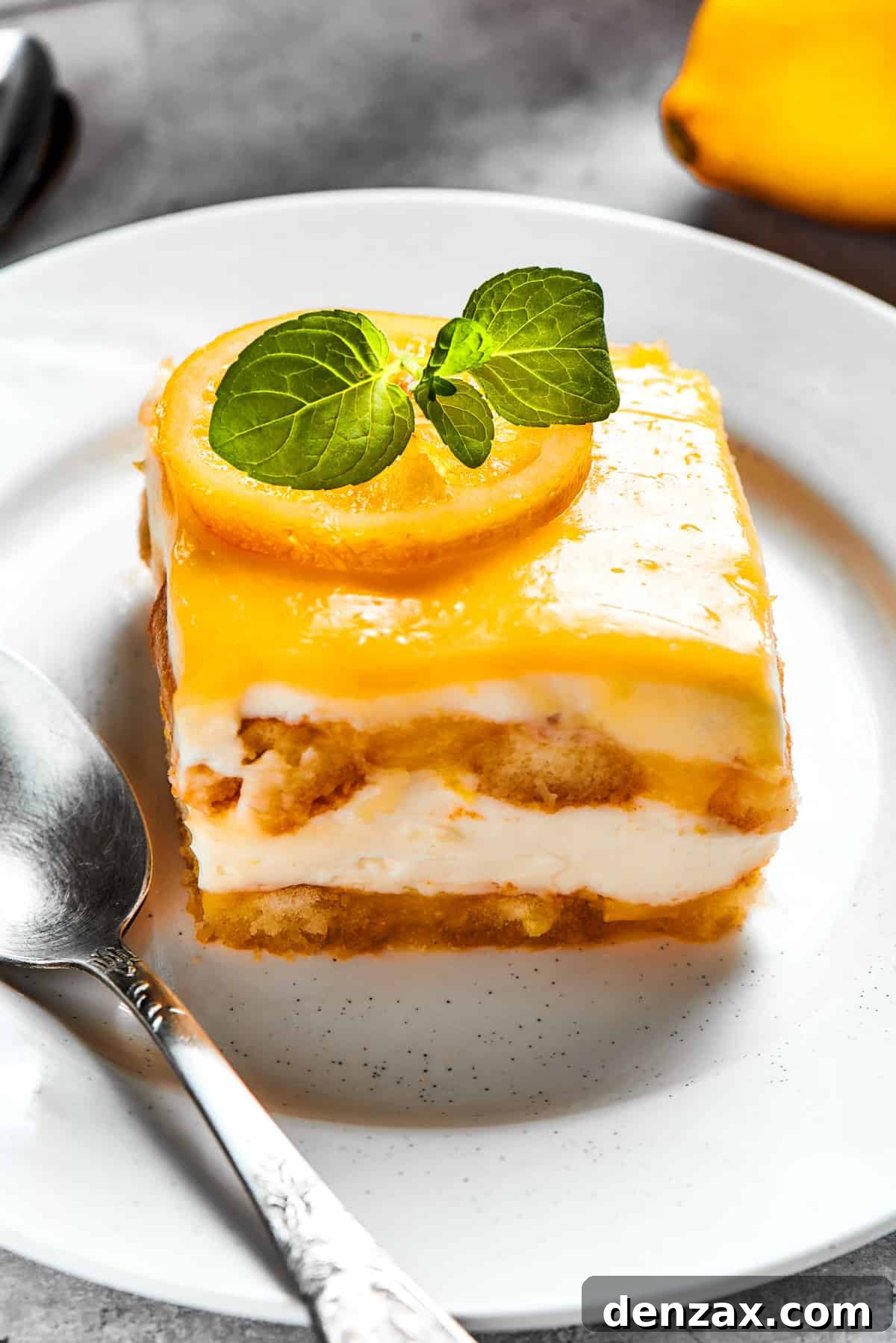 A perfectly square slice of lemon tiramisu rests on a white dessert plate, garnished with fresh basil or mint, showcasing its layered beauty.