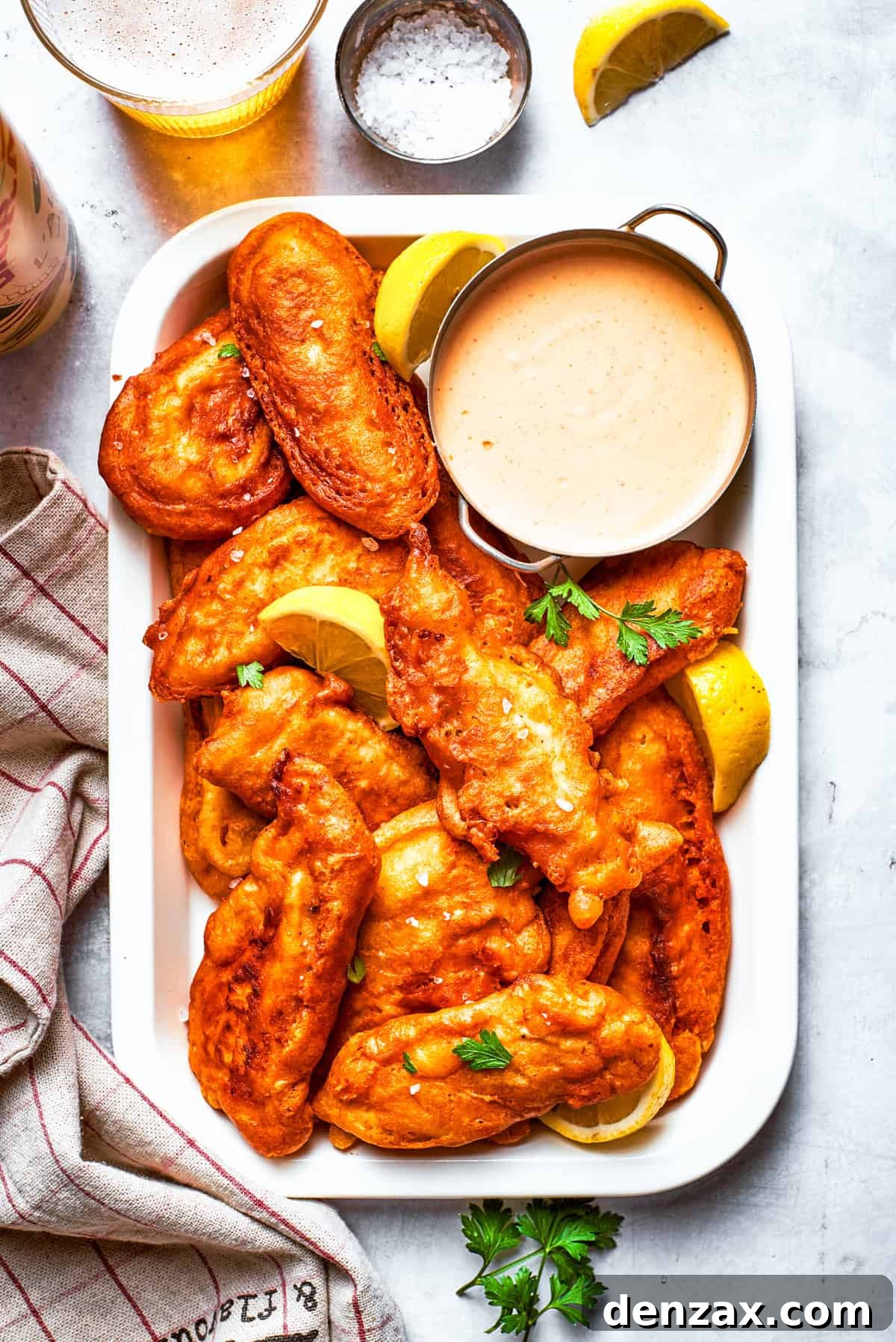 A rustic wooden platter features an enticing arrangement of golden-brown beer-battered chicken pieces, accompanied by a small, elegant bowl of creamy dipping sauce, ready to be enjoyed.