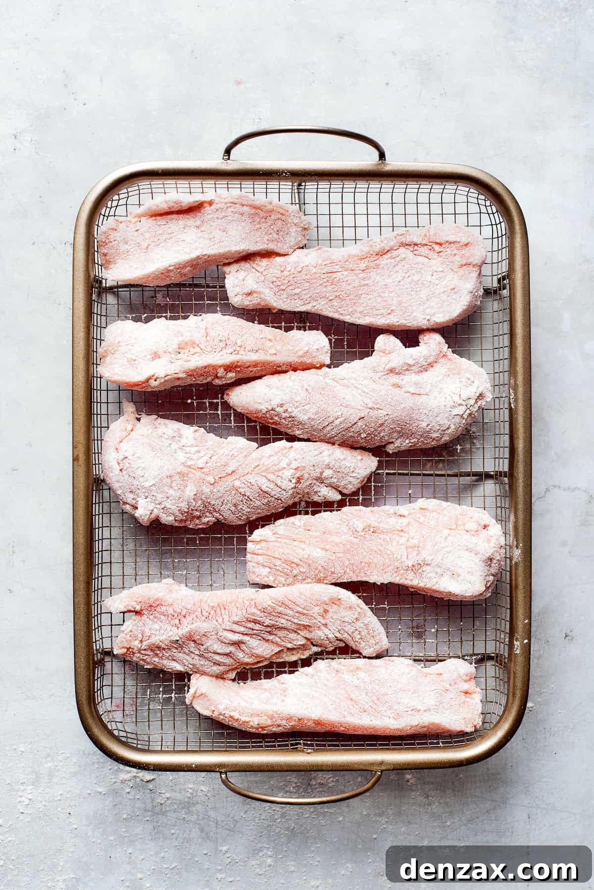 Uniformly sliced pieces of raw chicken breasts are neatly arranged on a wire rack, allowing for air circulation and ensuring proper seasoning and drying before coating.