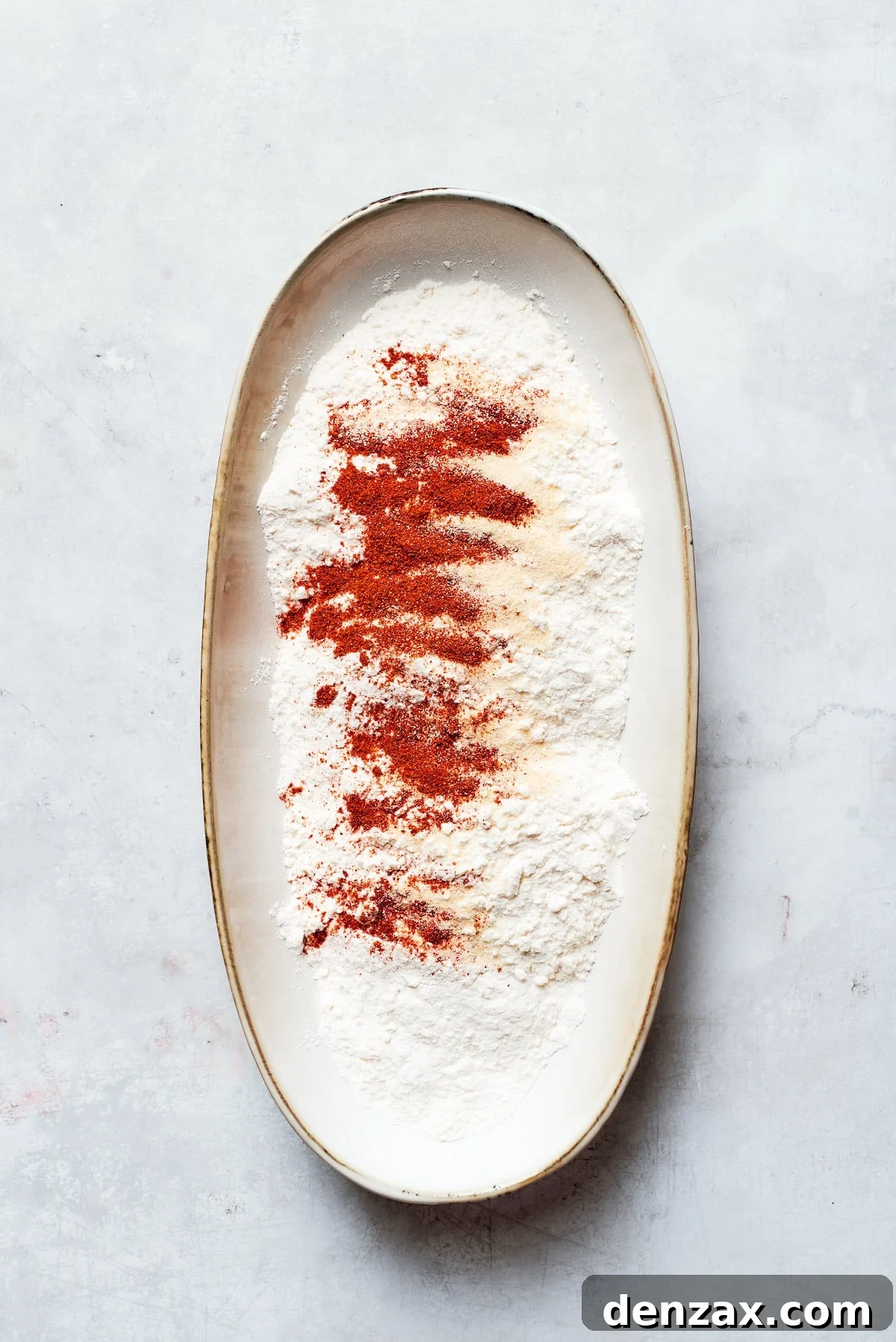 A close-up shot of dry ingredients for the dredging mixture being combined in a shallow bowl. All-purpose flour is mixed with various spices, ensuring an even distribution of flavor.