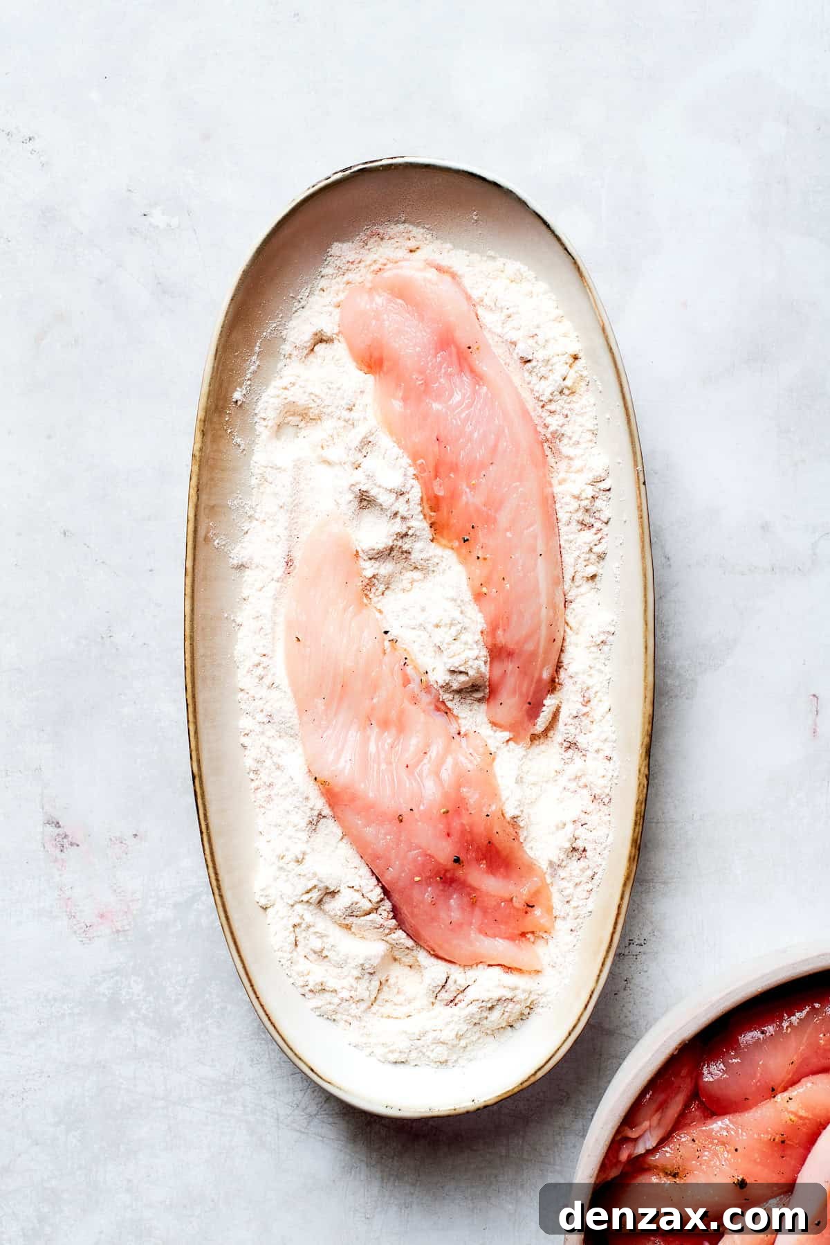 A hand gently dredging a chicken strip in a bowl of seasoned flour, ensuring a thorough and even coating on all sides before it is dipped into the wet batter.