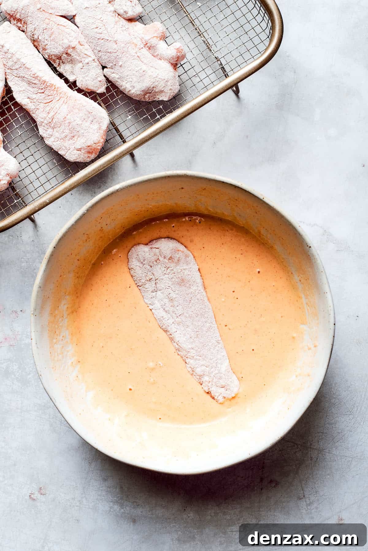 A piece of flour-dredged chicken is carefully submerged into a bowl of smooth, pale yellow beer batter, ensuring complete coverage for a perfectly crispy fried coating.