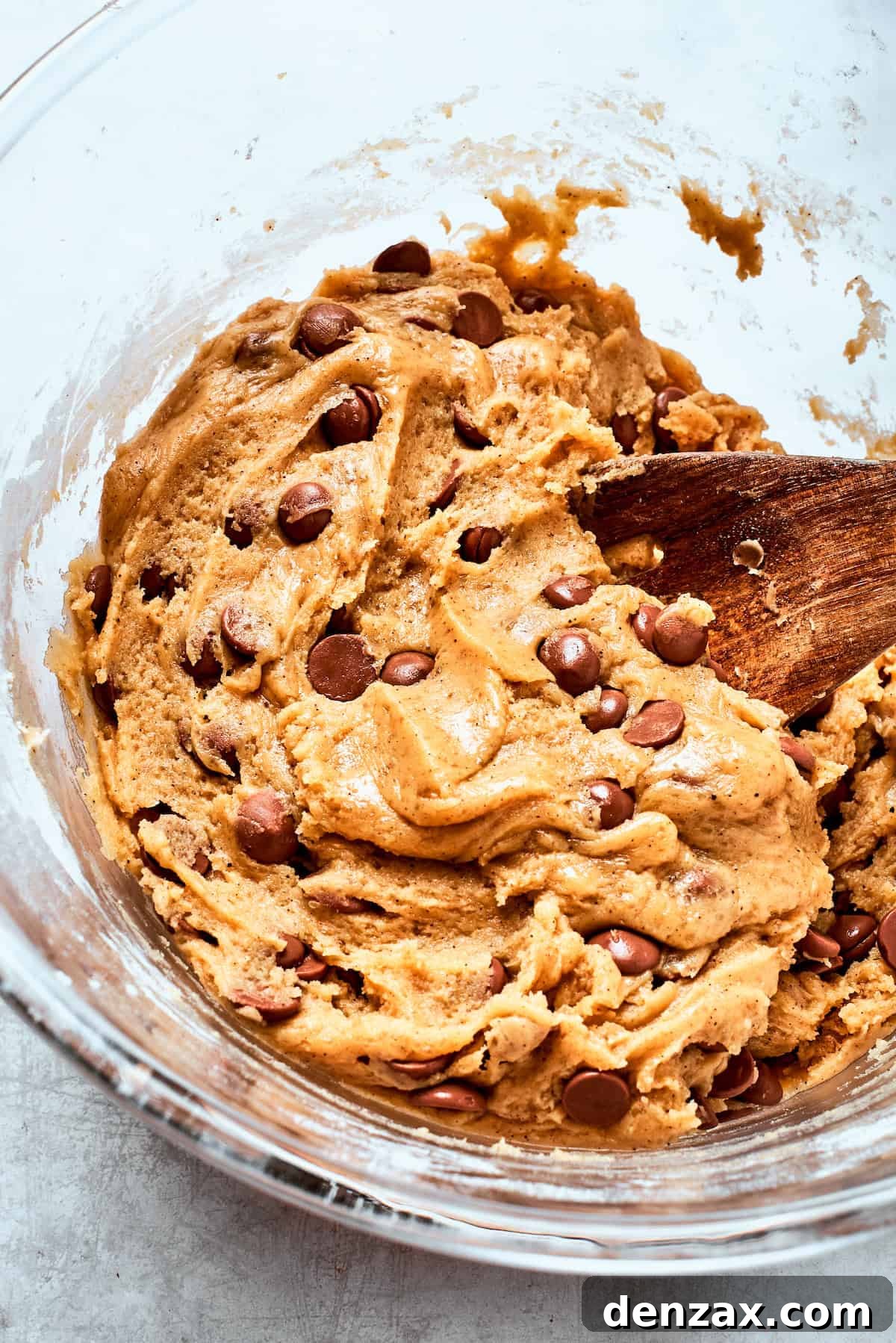 A wooden spoon gently stirs generous amounts of chocolate chips into the thick cookie dough.