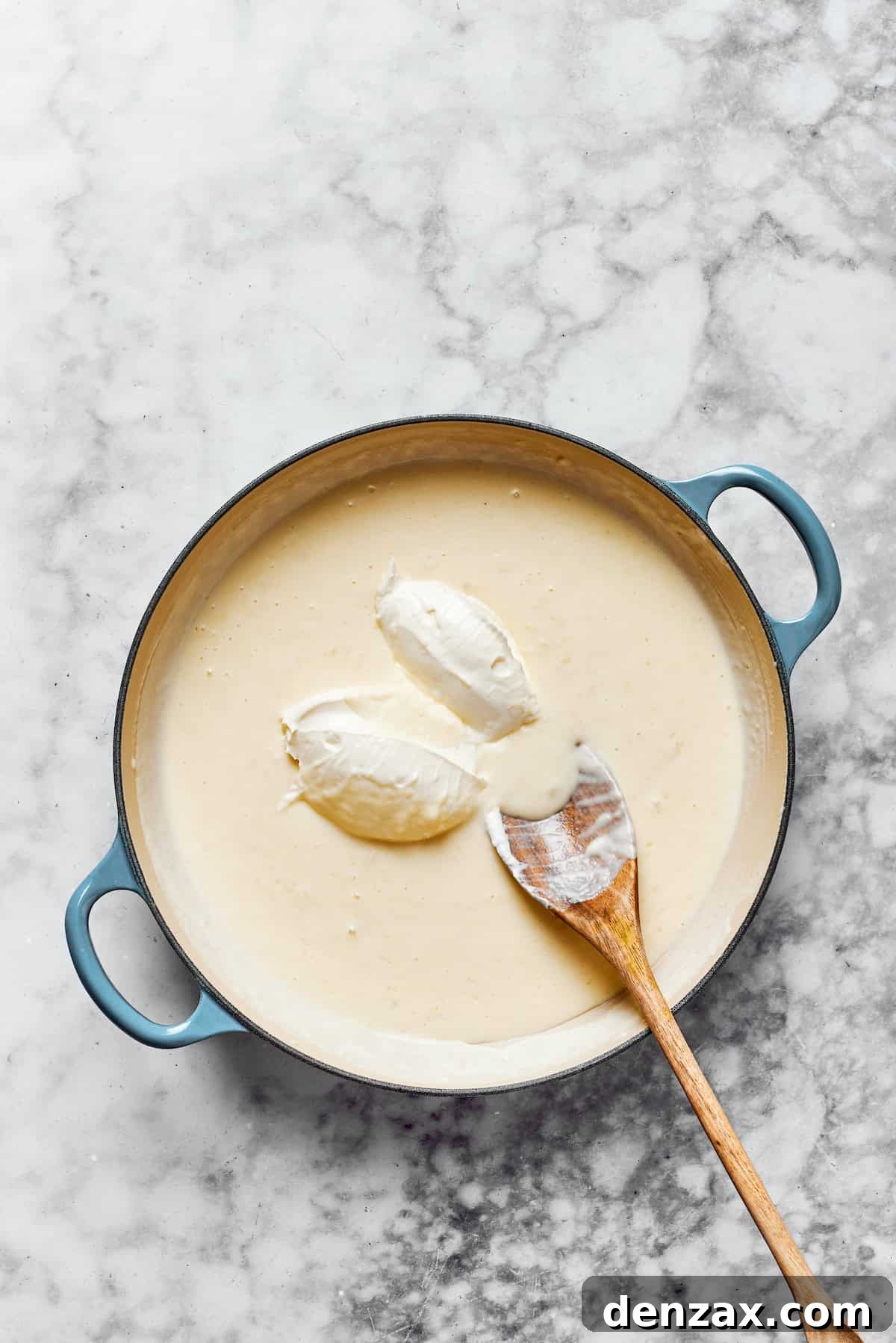 Cubed cream cheese being stirred into the creamy sauce in a pan, alongside Parmesan cheese.