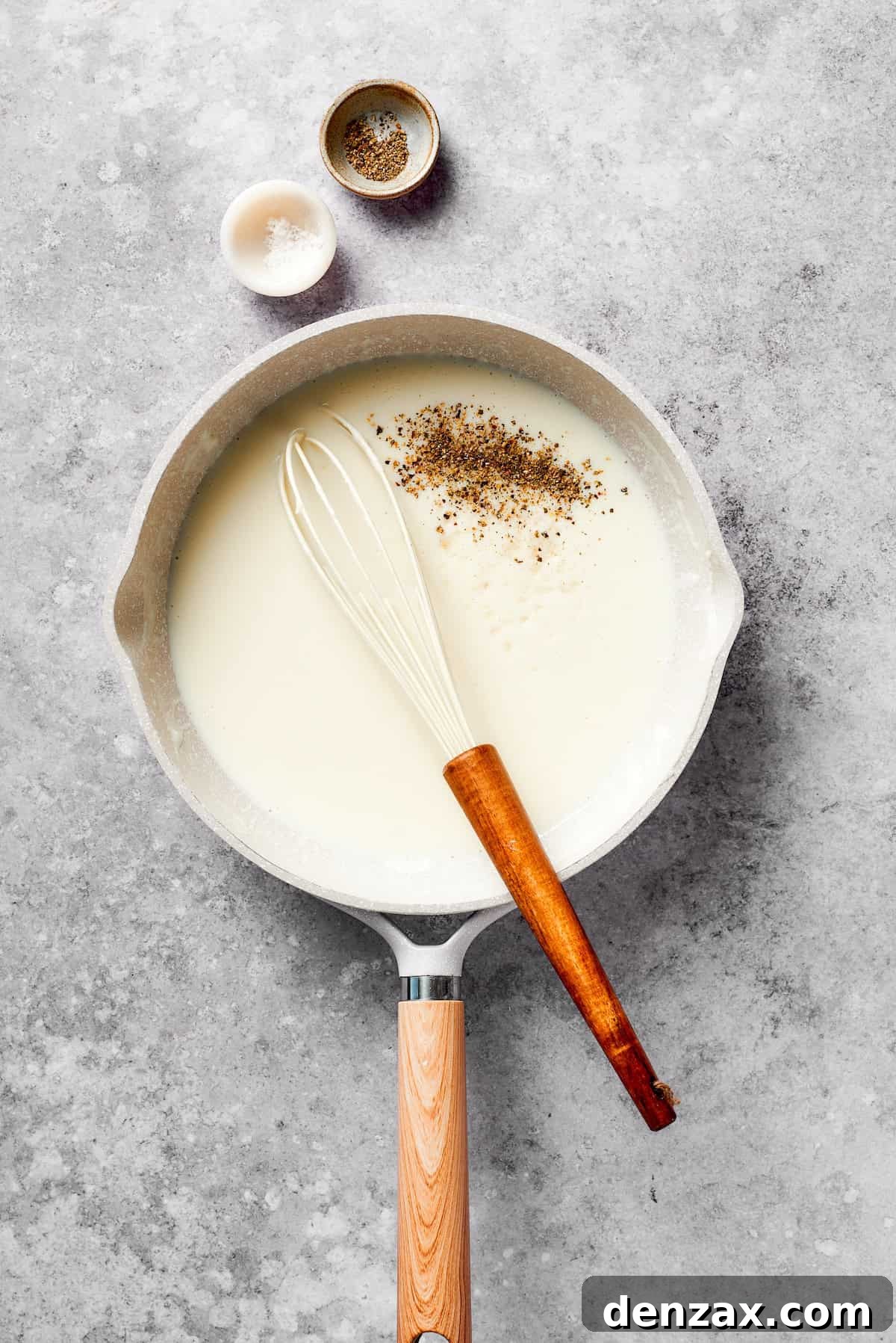 Milk being gradually whisked into the roux, along with a dash of pepper, creating a thickening béchamel sauce.