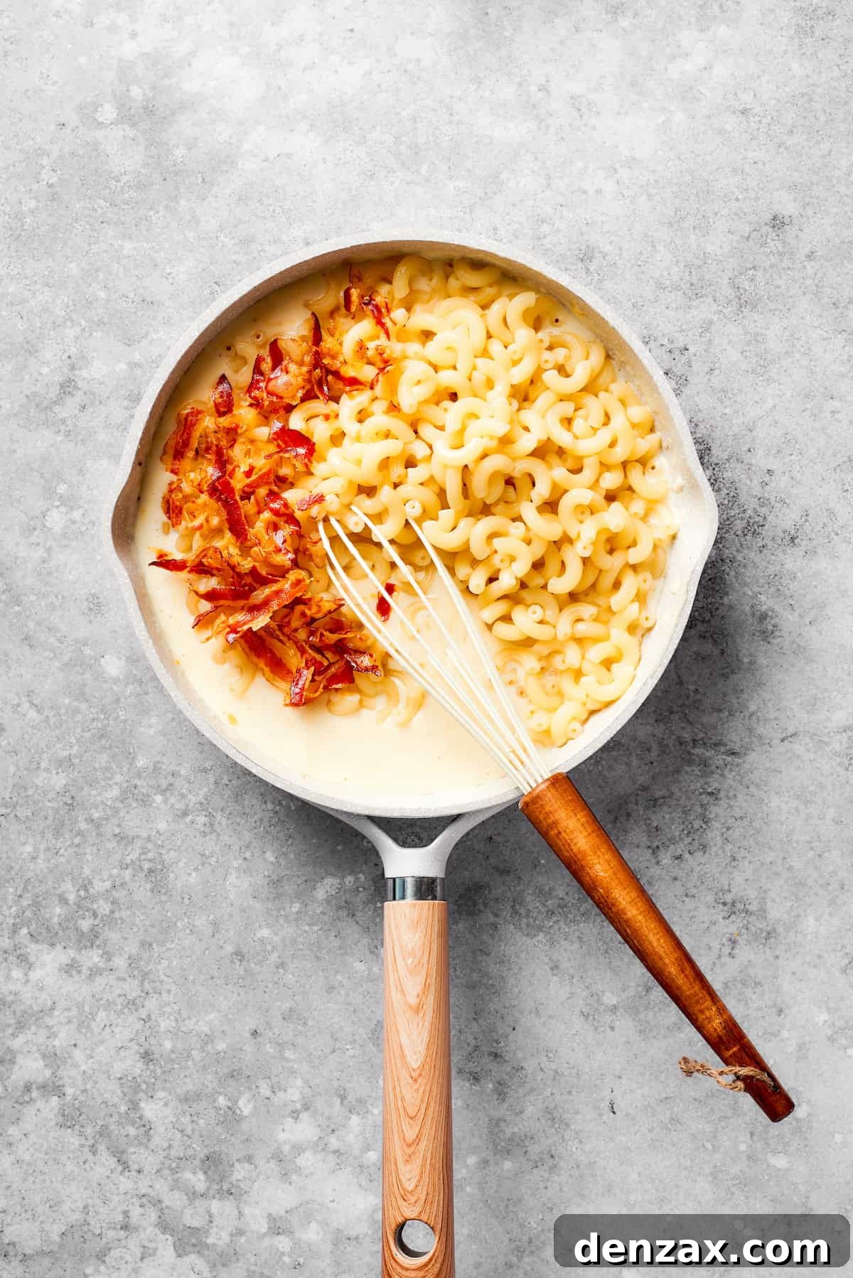Cooked macaroni and crispy bacon being added to the creamy, cheesy béchamel sauce in a large pan.