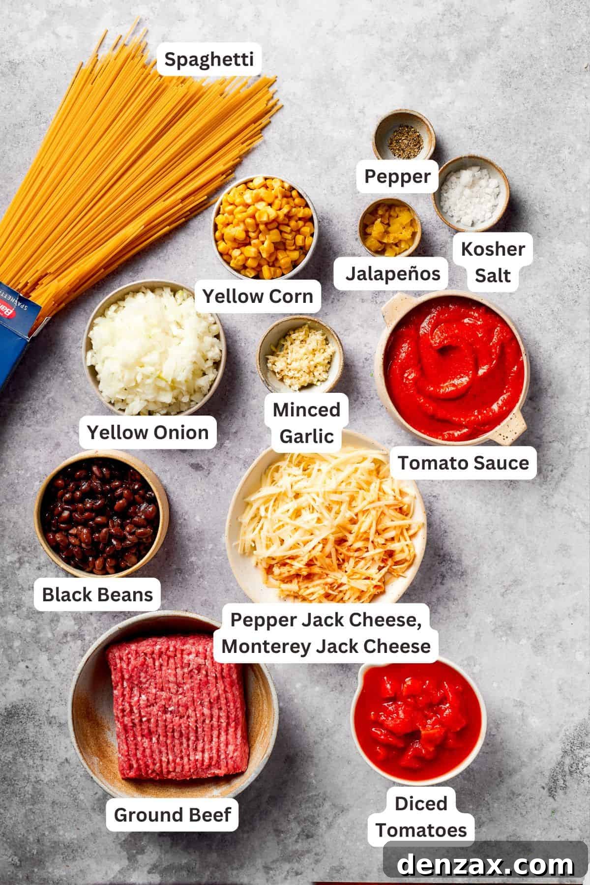 Individual ingredients for Mexican taco spaghetti are displayed, including uncooked spaghetti, diced onion, minced garlic, ground beef, canned tomato sauce, diced tomatoes, chopped jalapeños, corn, black beans, and shredded cheeses.