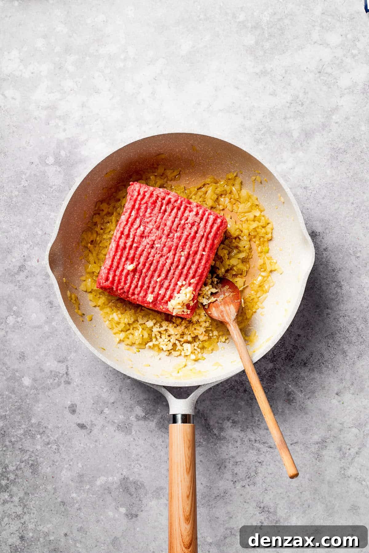 A block of ground beef being added to a large, hot frying pan with sautéing onions, ready to be cooked and browned.