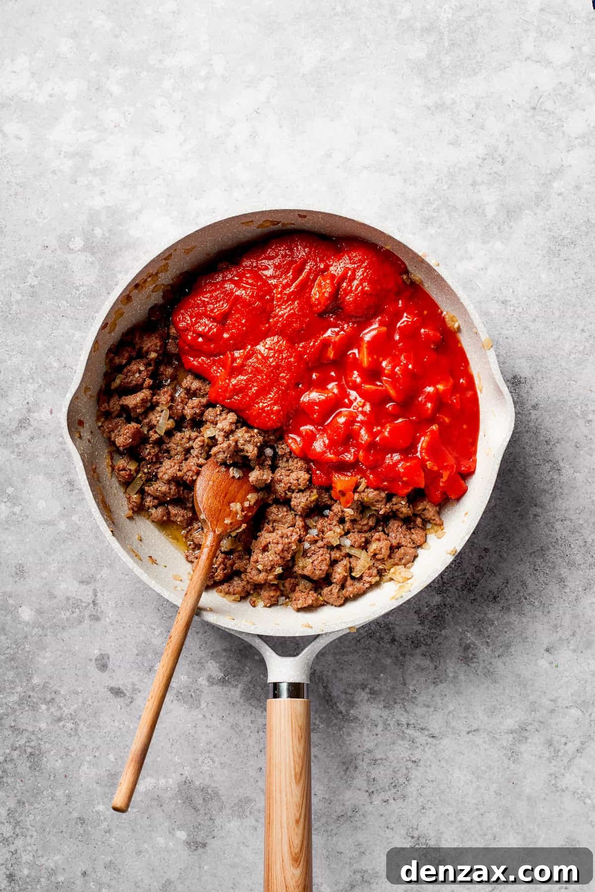 Canned diced tomatoes and tomato sauce being stirred into browned ground beef and onions in a frying pan, creating a rich sauce.