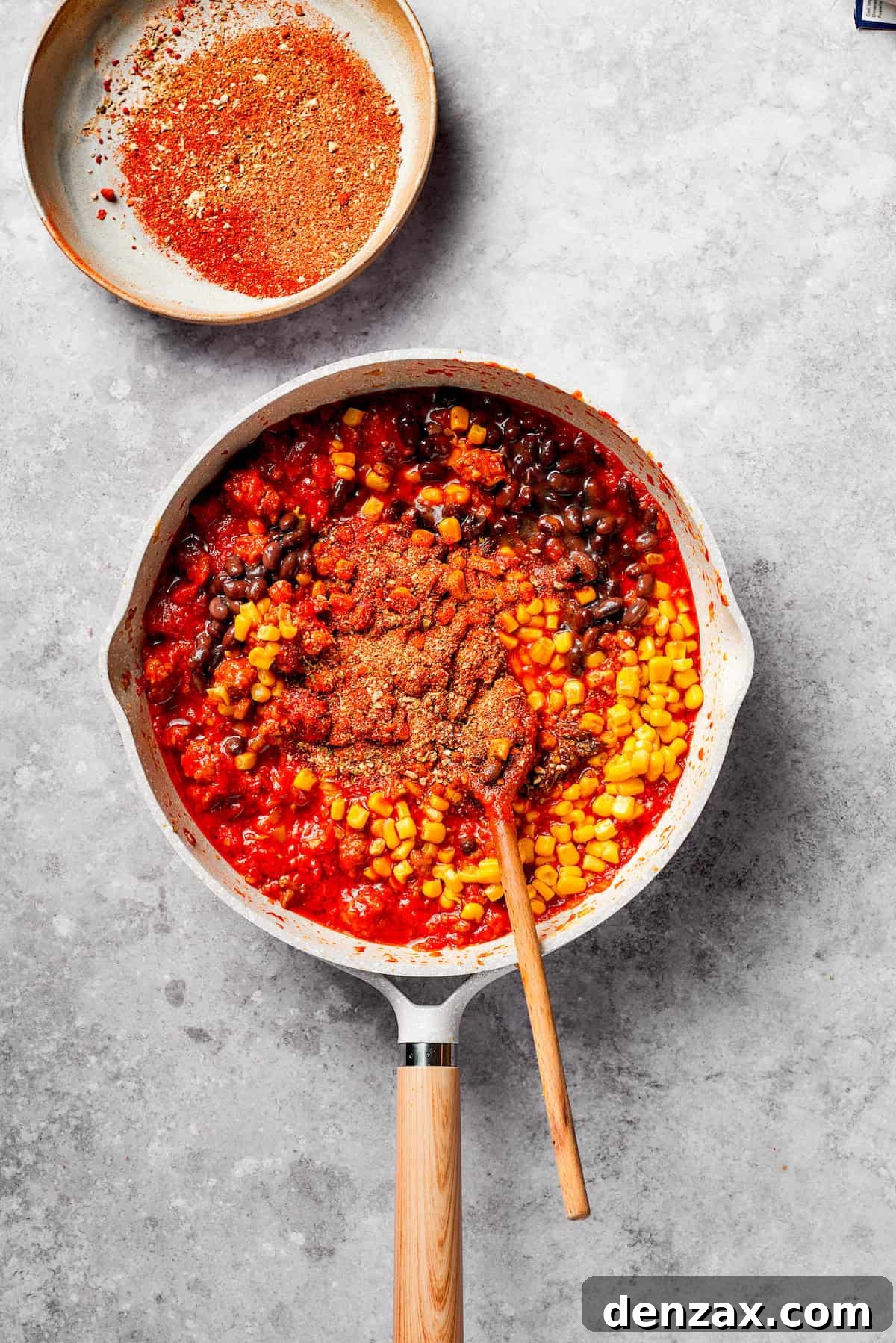 Black beans, corn, and chopped jalapeños being stirred into the simmering ground beef and tomato mixture in a large pan.