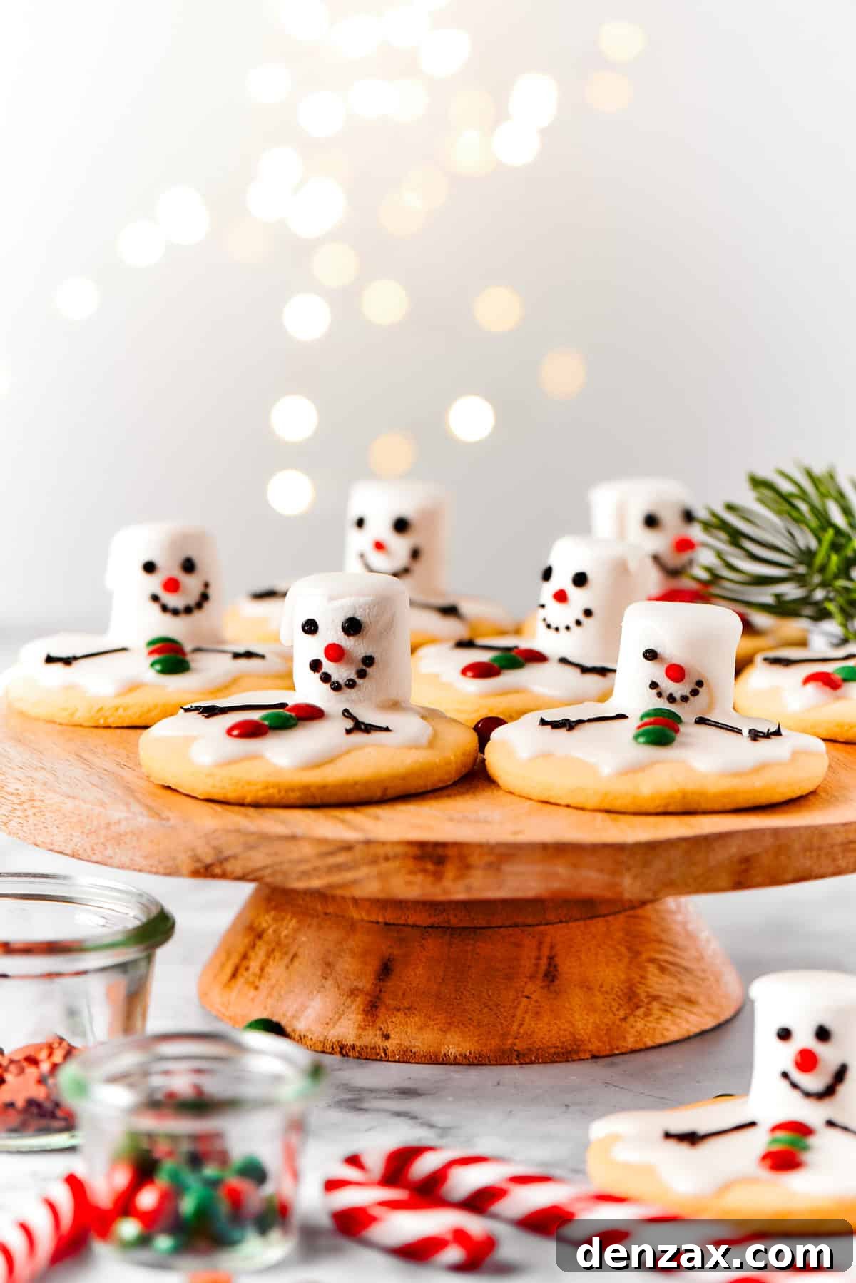The cutest Melted Snowman Cookies top buttery homemade sugar cookies with a thick vanilla frosting and a fluffy marshmallow! Melting snowman cookies are arranged on a wooden cake plate, surrounded by various holiday decorations like pinecones and ornaments, creating a festive scene.
